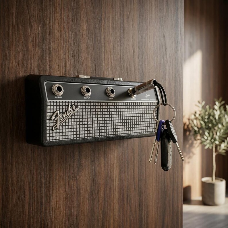 Puede incluir: Llavero negro con forma de amplificador Fender, montado en una puerta de madera. El llavero tiene detalles plateados y el logotipo de Fender. Las llaves cuelgan de una entrada jack de guitarra.