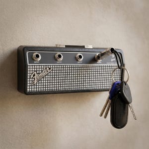 Puede incluir: Un llavero negro y plateado con forma de amplificador de guitarra, con el logotipo de Fender. El llavero tiene tres conectores de entrada y un enchufe plateado con un cable negro que sujeta llaves.
