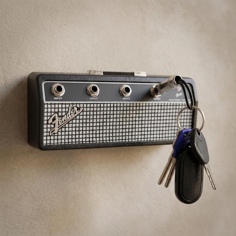 Puede incluir: Un llavero negro y plateado con forma de amplificador de guitarra, con el logotipo de Fender. El llavero tiene tres conectores de entrada y un enchufe plateado con un cable negro que sujeta llaves.