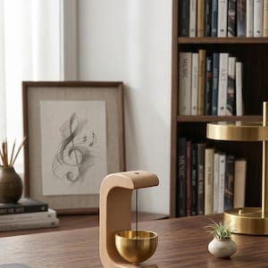 Puede incluir: Un porta incienso de madera y latón con un diseño curvo. El soporte presenta un pequeño cuenco de latón suspendido de un marco de madera. Un dibujo enmarcado de notas musicales y una estantería se encuentran en el fondo. El escritorio es de madera oscura.