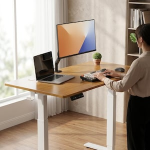 Puede incluir: Un escritorio regulable en altura con una tapa de madera marrón claro y patas blancas. Un monitor de ordenador negro, un portátil y un teclado están sobre el escritorio. Una persona está usando el teclado. Una pequeña planta en maceta está en el escritorio.