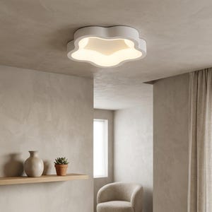 May include: A white, cloud-shaped ceiling light fixture illuminates a room. The fixture has a soft, organic design and is mounted on a neutral-colored ceiling. The room features a minimalist aesthetic with a shelf, vases, and a small potted plant.