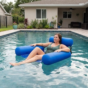 May include: A person relaxes in a blue inflatable pool chair in a swimming pool. The chair has a backrest and armrests. The person is wearing a green swimsuit. The pool is surrounded by a patio and a house.