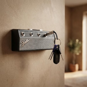 Puede incluir: Un llavero negro y plateado con forma de amplificador Fender montado en una pared. El llavero tiene cuatro conectores de entrada y un dise&ntilde;o de rejilla de altavoz. Un juego de llaves cuelga de uno de los conectores de entrada.