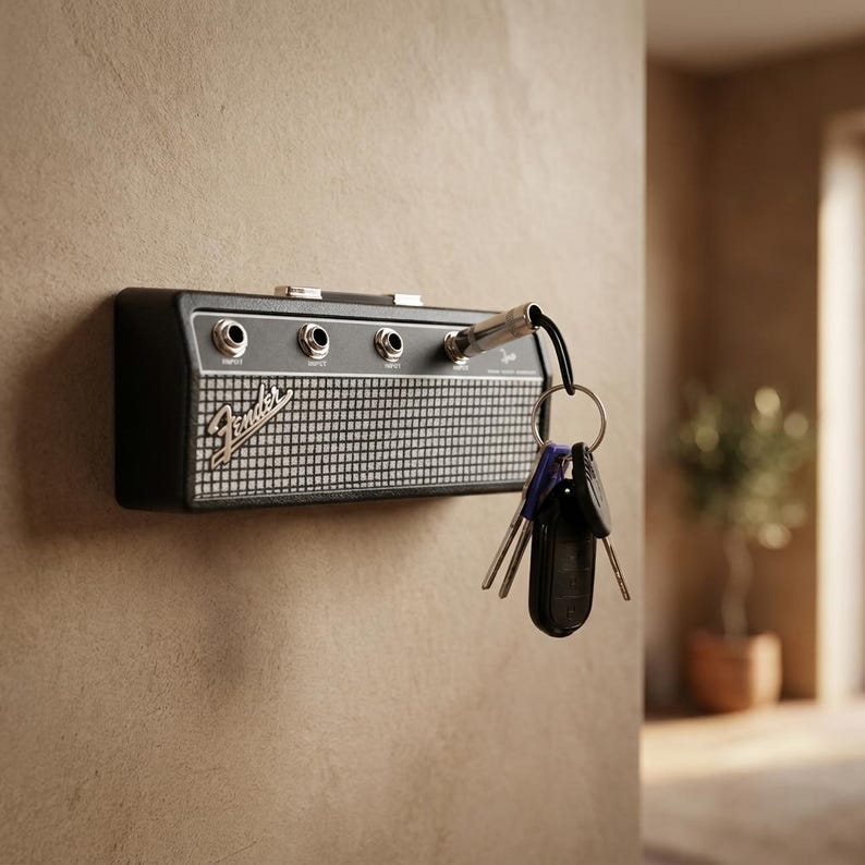 Puede incluir: Un llavero negro y plateado con forma de amplificador Fender montado en una pared. El llavero tiene cuatro conectores de entrada y un dise&ntilde;o de rejilla de altavoz. Un juego de llaves cuelga de uno de los conectores de entrada.