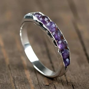 May include: A silver ring featuring six round, deep purple gemstones. The ring is displayed on a wooden surface, highlighting the contrast between the polished metal and the wood's natural grain.