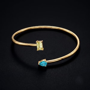 Puede incluir: Una pulsera abierta de oro con una piedra preciosa amarilla de corte esmeralda en un extremo y una piedra azul de corte de pera en el otro. La banda de metal tiene grabado el texto Always & Forever a lo largo de la curva interior pulida.