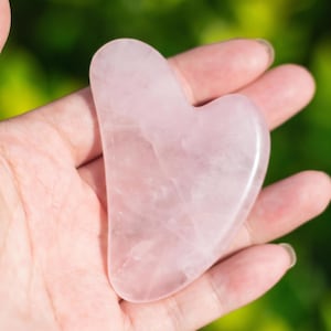 May include: A heart-shaped, light pink rose quartz gua sha tool. The smooth, polished stone is held in a hand against a blurred green background. The gua sha tool is a beauty and wellness product.
