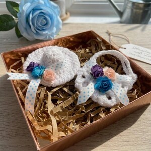 May include: A white crocheted hat and purse set, decorated with blue, purple, and peach-colored flowers and polka dot ribbon, presented in a copper-colored box with paper filler. A blue rose is in the background.