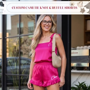 May include: Hot pink cami top and ruffle shorts set. The top has tie knot straps and the shorts have a ruffle hem. The set is personalized with the letter "C" on the top and the name "Cleo" on the shorts. The text "CUSTOM CAMI TIE KNOT & RUFFLE SHORTS" is at the top.