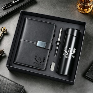 Può includere: Un set regalo nero contenente un taccuino rilegato in pelle con chiusura argentata, una penna, un bicchiere termico nero e una piccola gomma. Il taccuino e il bicchiere presentano un design a corna bianche con il nome "ROBERTS" e l'anno "EST. 2020".