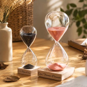 May include: Two glass hourglasses with wooden bases. The smaller hourglass contains black sand and is engraved with the text Love Beyond Time. The larger hourglass contains pink sand and is engraved with the text Our Forever Moment.
