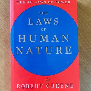 May include: A hardcover book titled The Laws of Human Nature by Robert Greene. The cover features a bright red background with a large blue circle in the center. The text is printed in white and gold, including the author name and book title.