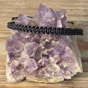 Puede incluir: Una pulsera de macramé negra descansa sobre un gran cristal de amatista. La pulsera está hecha de cordón negro tejido y tiene nudos corredizos ajustables. La amatista es de color púrpura intenso con formaciones cristalinas.