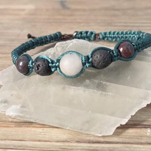 May include: A teal macrame bracelet featuring a combination of natural stone beads. The bracelet includes dark red, black, and white round beads. The bracelet is displayed on a white crystal against a wooden background.