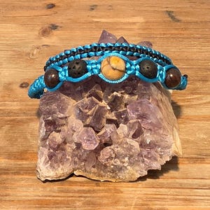 May include: Two macrame bracelets with natural stone beads. One bracelet is black, and the other is turquoise with brown, tan, and black beads. The bracelets are displayed on a purple crystal geode, set against a wooden background.