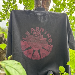 May include: A charcoal grey short-sleeve t-shirt featuring a circular maroon graphic on the back. The design includes a palm tree, swirling line patterns, and the text "LEARNING HUMAN" arranged in a circular border around the central illustration.