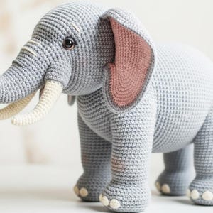 May include: A crocheted elephant toy in shades of gray, with ivory tusks and pink inner ears. The elephant has a textured surface and is standing on four legs. A charming handmade animal for children.