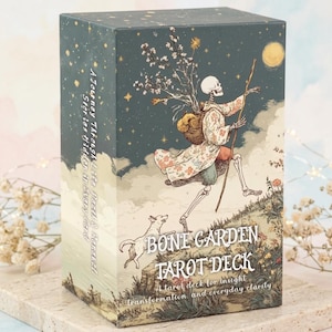 Op de afbeelding: Een rechthoekige tarotkaartendoos met de titel Bone Garden Tarot Deck. De voorkant toont een skelet dat wandelt met een staf en een bloemrijke rugzak, vergezeld door een hond onder een sterrenhemel. De tekst op de zijkant luidt A Journey Through Life, Death & Rebirth.