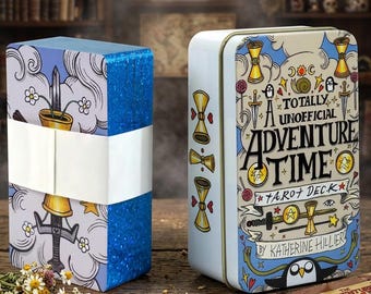 Op de afbeelding: Een tarotkaartspel met blauwe glitterranden verpakt in een witte papieren band, naast een rechthoekig wit metalen blik met de tekst A TOTALLY UNOFFICIAL ADVENTURE TIME TAROT DECK BY KATHERINE HILLIER en kleurrijke cartoonillustraties.