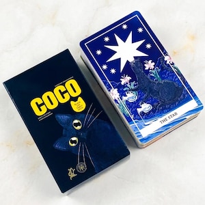 May include: A black rectangular box labeled COCO with a black cat illustration and a deck of tarot cards featuring a blue card titled THE STAR, which depicts a black cat under a large white star and smaller stars against a deep blue background.