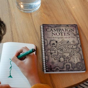 May include: A spiral-bound notebook with the words "CAMPAIGN NOTES" and "They Will Ignore All Of This" printed on the cover. The cover features a map design. A person is drawing in a lined notebook with a green pen.