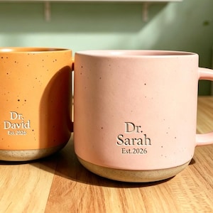 Puede incluir: Dos tazas de cerámica con acabado moteado, una naranja y otra rosa claro. Cada taza tiene un asa y una base de color claro. La taza naranja está grabada con Dr. David Est. 2026 y la taza rosa con Dr. Sarah Est. 2026.