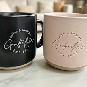 Puede incluir: Dos tazas de cerámica, una negra y otra rosa, con un diseño de Padrino y Madrina. La taza negra tiene "Colton & Emmett's Godfather EST. 2020" en blanco. La taza rosa tiene "Colton & Emmett's Godmother EST. 2026".