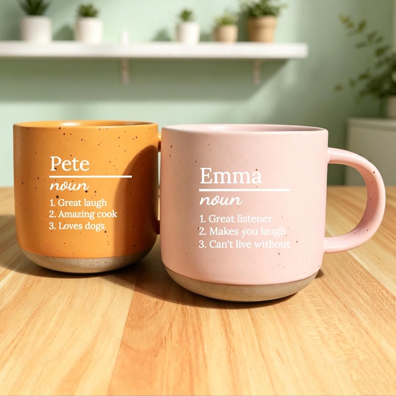 May include: Two ceramic mugs, one orange and one pink, sit side-by-side on a wooden surface. The orange mug has the name "Pete" and the pink mug has the name "Emma" printed on them, along with a list of positive attributes.