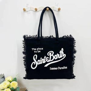 Puede incluir: Bolso tote negro con asas largas y bordes deshilachados. La bolsa presenta el texto "The place to be Saint Barth Summer Paradise" en escritura blanca. La bolsa est&aacute; colgada en una pared blanca.