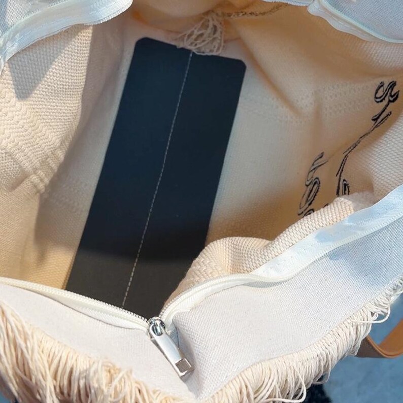 Pu&ograve; includere: Borsa tote color crema con chiusura a zip e dettagli con frange. L'interno della borsa presenta un'etichetta nera e un disegno stampato in nero. La borsa &egrave; realizzata in un materiale morbido e tessuto.