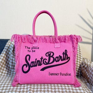 Puede incluir: Un bolso tote rosa brillante con asas y flecos a juego. El bolso presenta el texto "The place to be Saint Barth Summer Paradise" en escritura negra. El bolso est&aacute; hecho de un material suave.