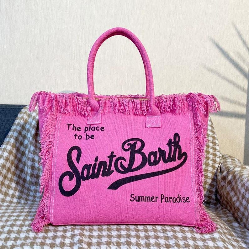 Puede incluir: Un bolso tote rosa brillante con asas y flecos a juego. El bolso presenta el texto "The place to be Saint Barth Summer Paradise" en escritura negra. El bolso est&aacute; hecho de un material suave.