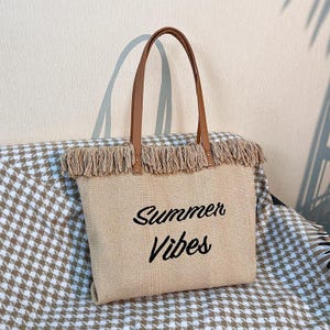 Pu&ograve; includere: Borsa tote beige con manici in pelle marrone e finiture con frange. Le parole "Summer Vibes" sono ricamate in nero sul davanti. La borsa &egrave; realizzata in materiale tessuto ed &egrave; progettata per trasportare oggetti.