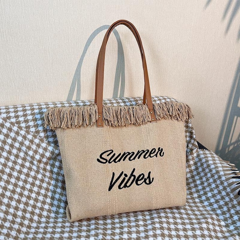Pu&ograve; includere: Borsa tote beige con manici in pelle marrone e finiture con frange. Le parole "Summer Vibes" sono ricamate in nero sul davanti. La borsa &egrave; realizzata in materiale tessuto ed &egrave; progettata per trasportare oggetti.