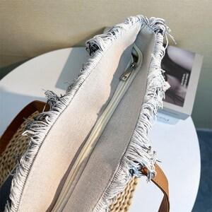 Pu&ograve; includere: Primo piano di una borsa tote beige con chiusura a cerniera. La borsa ha un dettaglio bordo sfilacciato con manici in pelle marrone. L'interno &egrave; di colore beige chiaro. La borsa &egrave; appoggiata su una superficie tessuta.