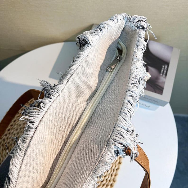 Pu&ograve; includere: Primo piano di una borsa tote beige con chiusura a cerniera. La borsa ha un dettaglio bordo sfilacciato con manici in pelle marrone. L'interno &egrave; di colore beige chiaro. La borsa &egrave; appoggiata su una superficie tessuta.