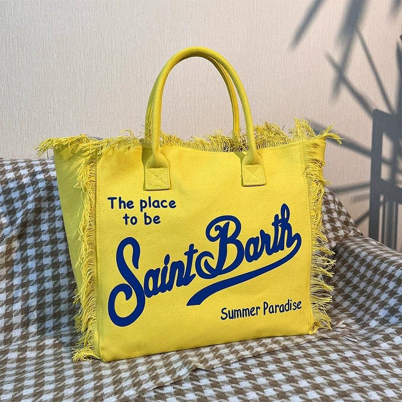 Puede incluir: Bolso tote amarillo con ribete de flecos y asas a juego. El bolso presenta el texto "The place to be Saint Barth Summer Paradise" en escritura azul. El bolso est&aacute; sobre un estampado de cuadros marrones y blancos.