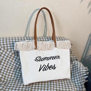 Pu&ograve; includere: Borsa tote in tela color crema con manici in pelle marrone. La borsa presenta un bordo con frange sulla parte superiore e le parole "Summer Vibes" in corsivo nero. La borsa &egrave; su una coperta fantasia.