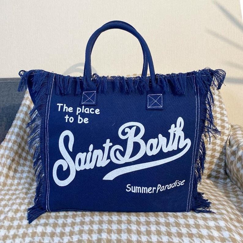 Puede incluir: Un bolso tote azul marino con texto blanco que dice "The place to be Saint Barth Summer Paradise". El bolso tiene dos asas y est&aacute; adornado con flecos.