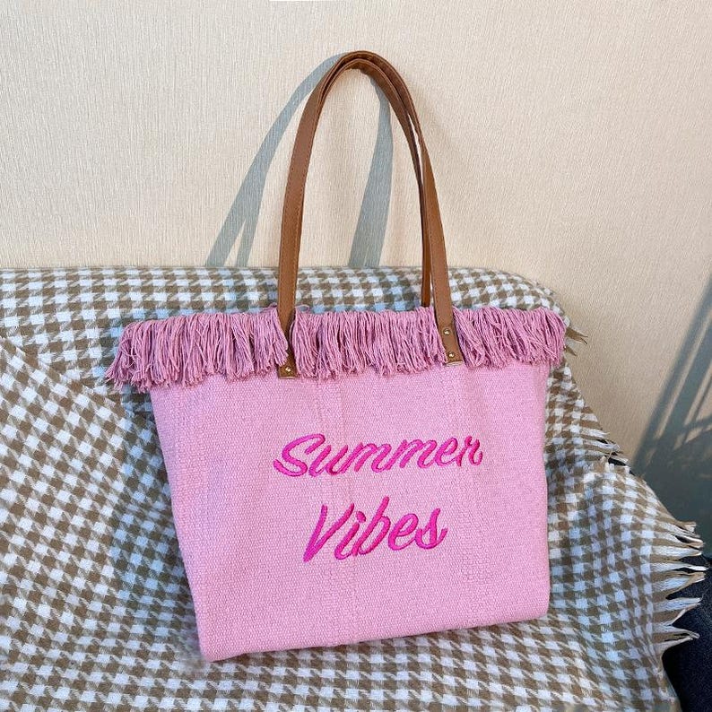 Pu&ograve; includere: Borsa tote rosa con manici in pelle marrone e finiture con frange. La borsa presenta la scritta ricamata "Summer Vibes" in rosa. La borsa &egrave; realizzata in tessuto.