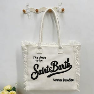 Puede incluir: Bolso tote blanco con flecos y dos asas. El bolso presenta el texto "The place to be Saint Barth Summer Paradise" en escritura negra. El bolso est&aacute; colgado en una pared blanca.