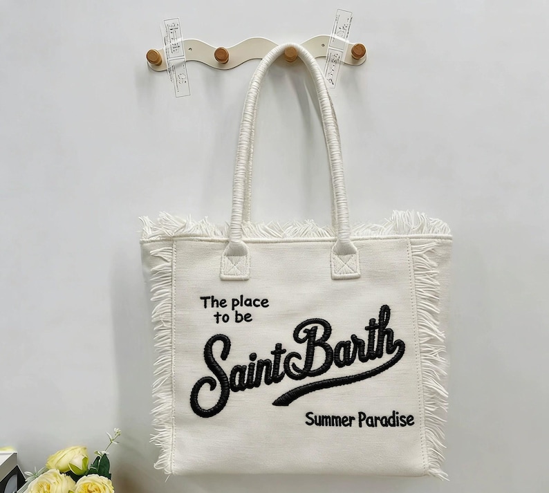 Puede incluir: Bolso tote blanco con flecos y dos asas. El bolso presenta el texto "The place to be Saint Barth Summer Paradise" en escritura negra. El bolso est&aacute; colgado en una pared blanca.