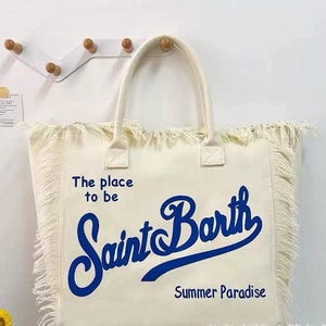 Puede incluir: Bolso tote de color crema con flecos y asas. El bolso presenta el texto "The place to be Saint Barth Summer Paradise" en escritura azul. El bolso est&aacute; hecho de un material similar a la lona.