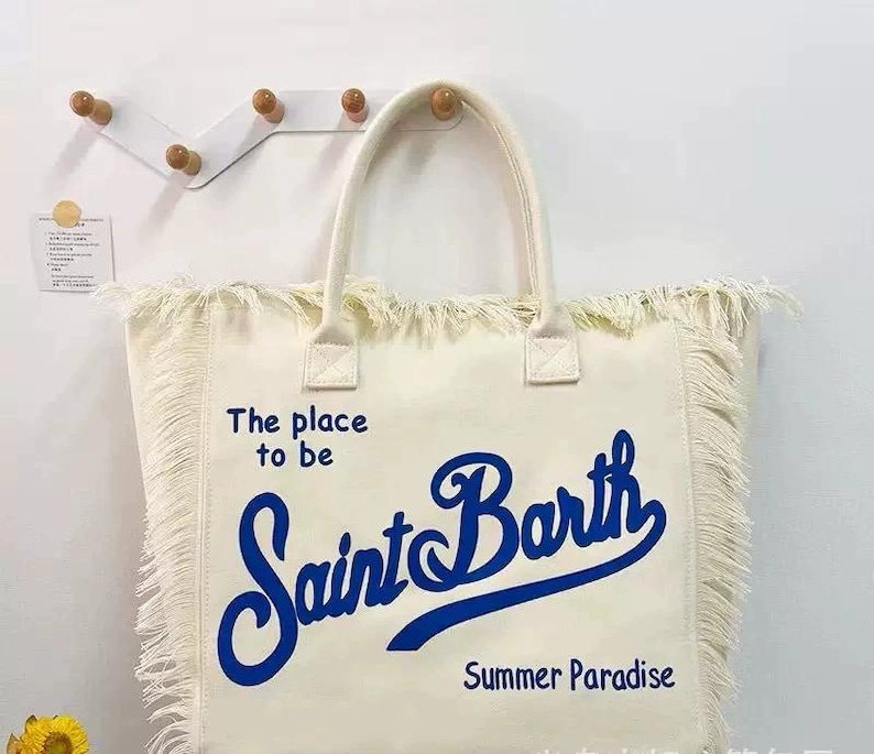 Puede incluir: Bolso tote de color crema con flecos y asas. El bolso presenta el texto "The place to be Saint Barth Summer Paradise" en escritura azul. El bolso est&aacute; hecho de un material similar a la lona.