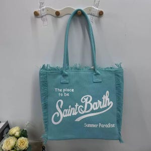 Puede incluir: Un bolso tote turquesa con asas largas y flecos. El bolso presenta el texto "The place to be Saint Barth Summer Paradise" en escritura blanca. El bolso est&aacute; colgado en una pared blanca.