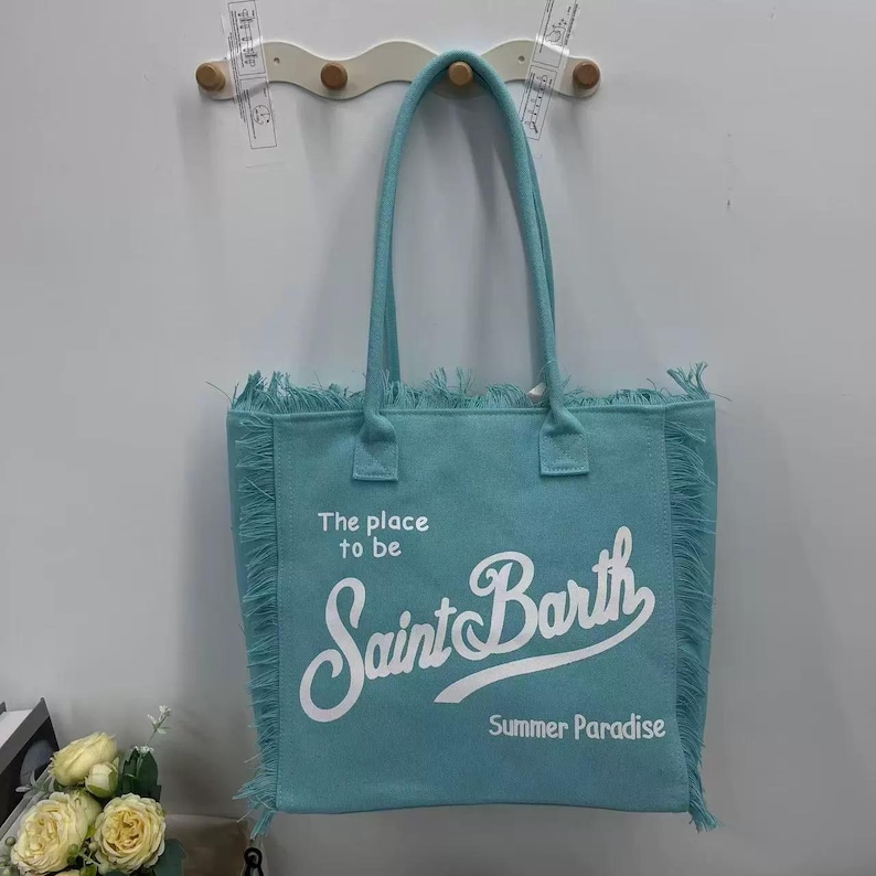 Puede incluir: Un bolso tote turquesa con asas largas y flecos. El bolso presenta el texto "The place to be Saint Barth Summer Paradise" en escritura blanca. El bolso est&aacute; colgado en una pared blanca.