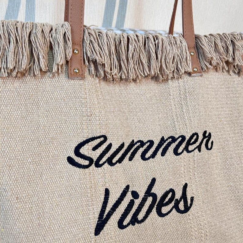 Pu&ograve; includere: Borsa tote beige con manici in pelle marrone e finiture con frange. Le parole "Summer Vibes" sono ricamate in nero sul davanti. La borsa &egrave; realizzata in tessuto intrecciato.