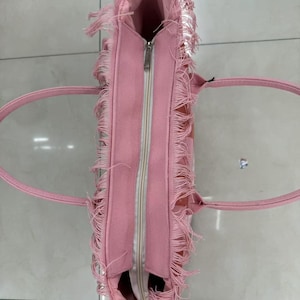 Puede incluir: Bolso tote rosa con cierre de cremallera y detalles de flecos. El bolso tiene dos asas y forma rectangular. El interior es visible y parece ser de un tono rosa m&aacute;s claro. El bolso est&aacute; hecho de un material suave.