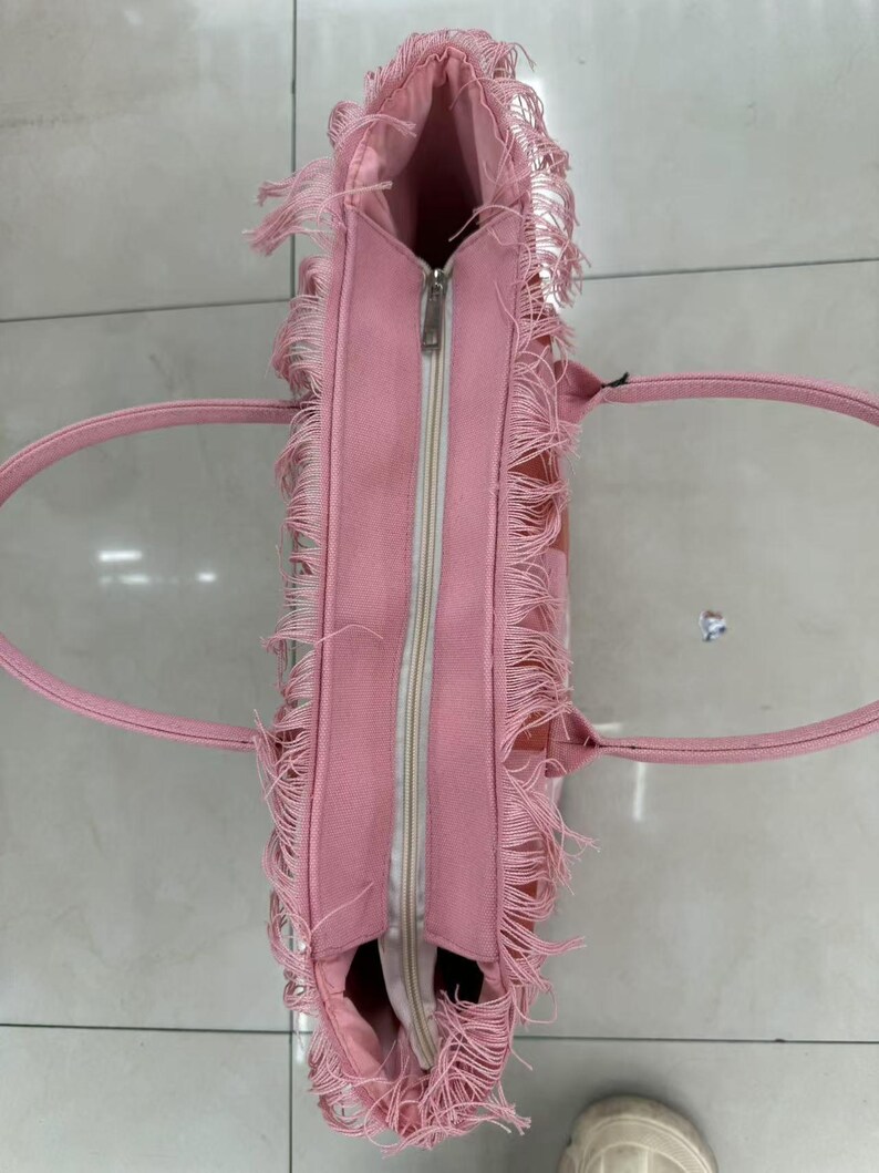 Puede incluir: Bolso tote rosa con cierre de cremallera y detalles de flecos. El bolso tiene dos asas y forma rectangular. El interior es visible y parece ser de un tono rosa m&aacute;s claro. El bolso est&aacute; hecho de un material suave.